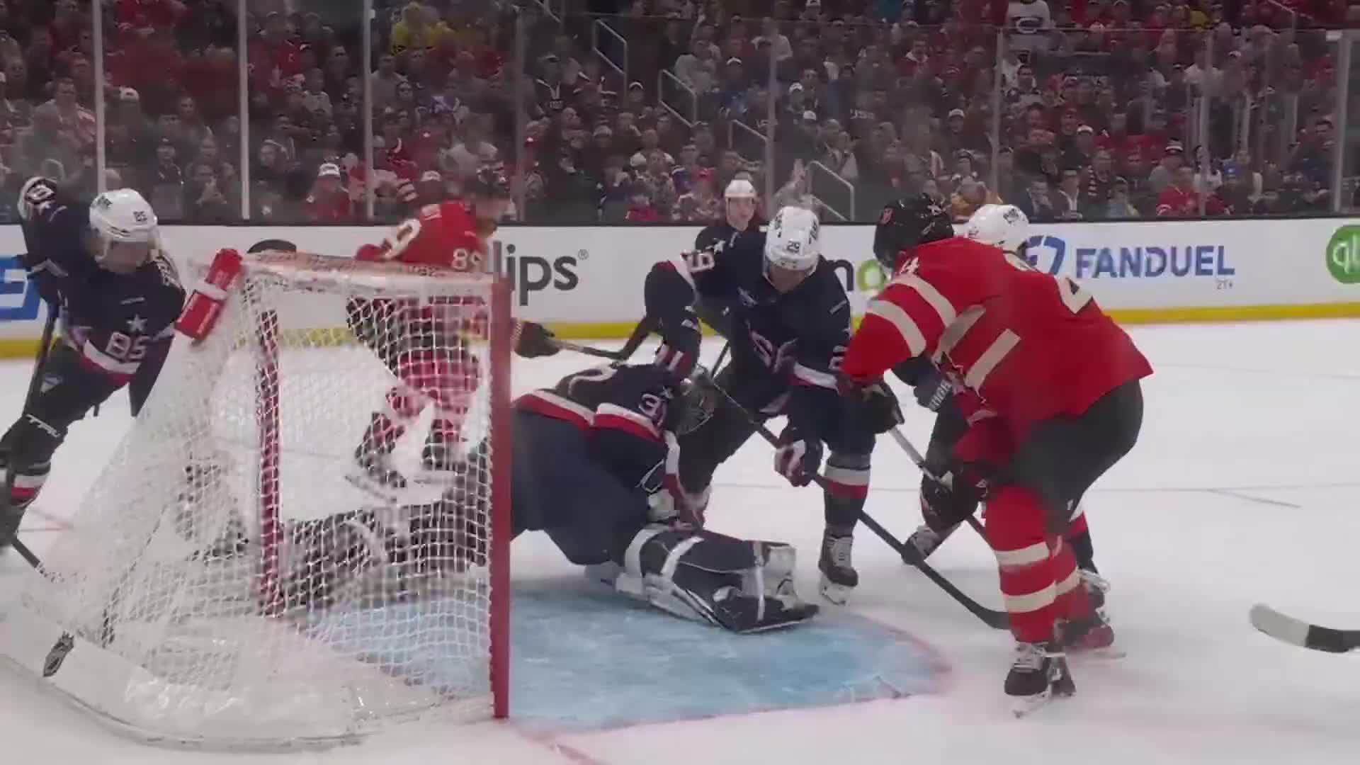 A goalie in red sprawls across the crease, his pads outstretched as players in blue and red jerseys converge. The puck is somewhere in the scramble, the crowd a blur of anticipation behind the boards.