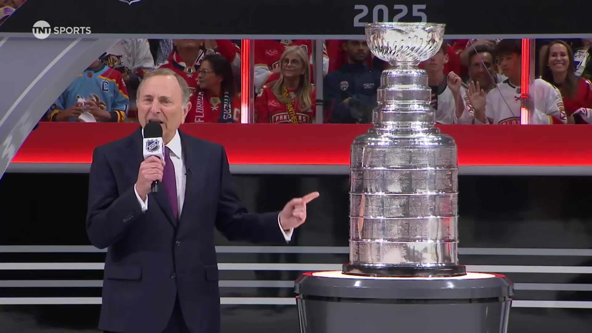 The commissioner stands at a podium, microphone in hand, gesturing towards the Stanley Cup. A crowd watches from the stands behind a red railing.