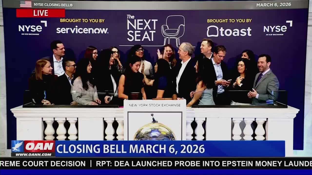 A group of people stand on a platform at the New York Stock Exchange, some smiling and looking at phones. The OANN chyron below announces the closing bell for March 6, 2026.