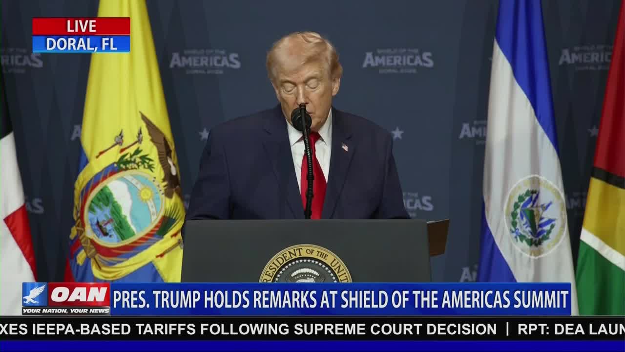 President Trump stands at a podium, speaking at the Shield of the Americas Summit in Doral, Florida. Flags from various nations stand behind him.