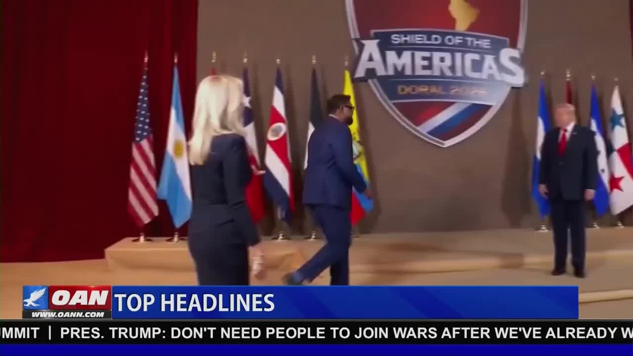 A woman in a dark suit walks past the American flag and other national banners. A man in a blue suit strides by, heading towards a large shield graphic behind him. President Trump stands to the right, facing forward.