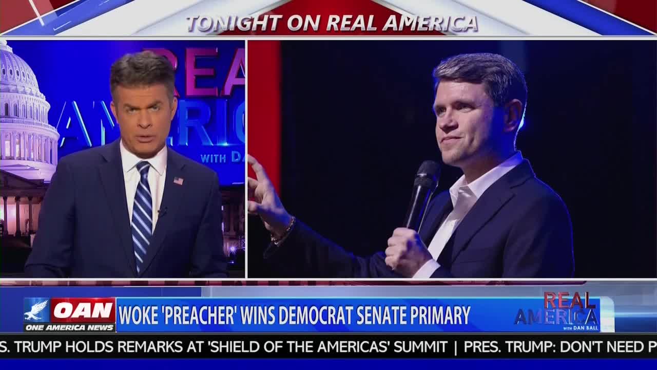A man in a dark suit and white shirt speaks into a microphone, gesturing with his left hand. Beside him, a news anchor in a blue suit and striped tie looks directly ahead. A banner at the bottom of the screen reads "OAN WOKE 'PREACHER' WINS DEMOCRAT SENATE PRIMARY."