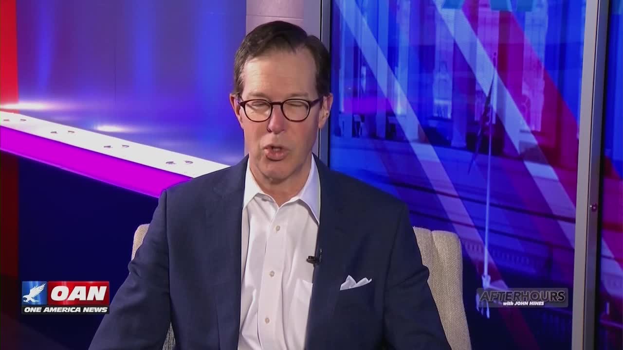 A man in a suit speaks into a microphone on the set of One America News. The backdrop behind him features a patriotic motif with red and blue stripes.