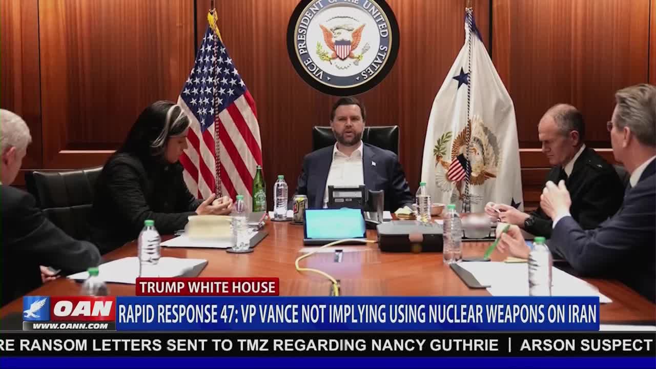 A group of people are seated around a large wooden table in a room with wood-paneled walls. An American flag stands to the left of a man in a suit, who is speaking. To his right, an official in a military uniform looks down at his hands.