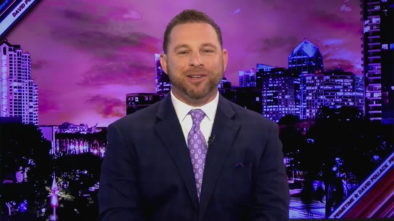A man in a dark suit and a patterned tie stands before a cityscape bathed in purple light. The backdrop features tall buildings with illuminated windows, suggesting a night scene in a major American city.