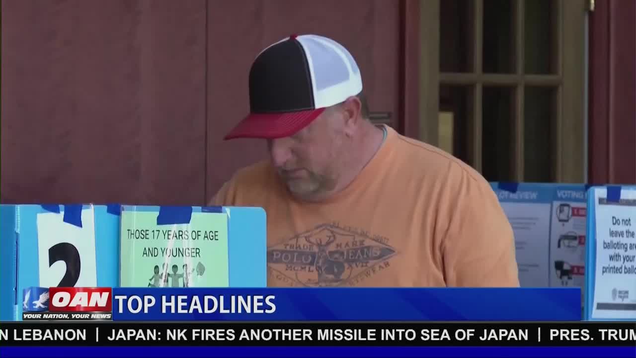 A man in an orange shirt and a red and white trucker hat leans over a table, focused on something out of frame. Blue dividers with signs are set up in front of him, one displaying "THOSE 17 YEARS OF AGE AND YOUNGER."