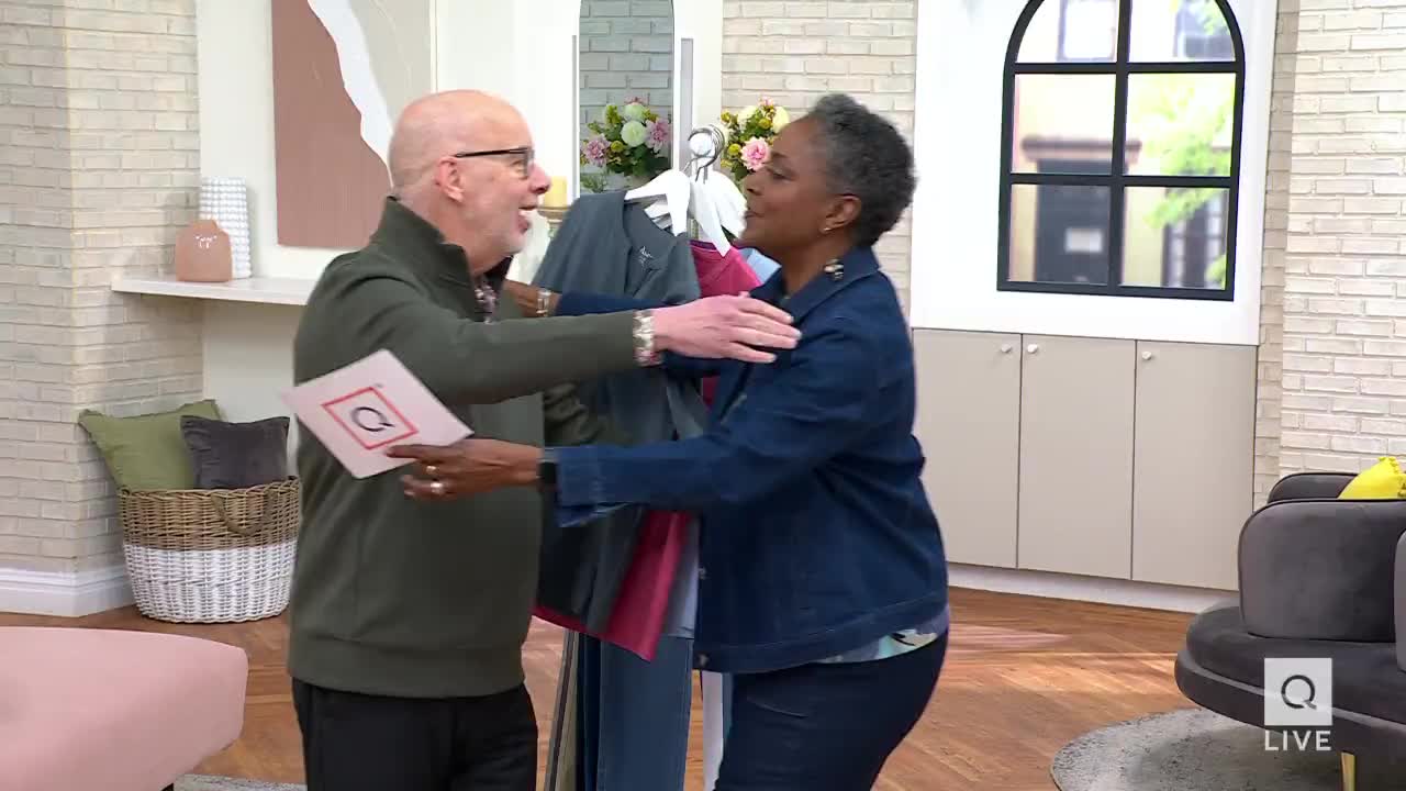 A man in a green sweater holds up a QVC card as he embraces a woman in a denim jacket. They stand in a brightly lit studio with clothing racks behind them.