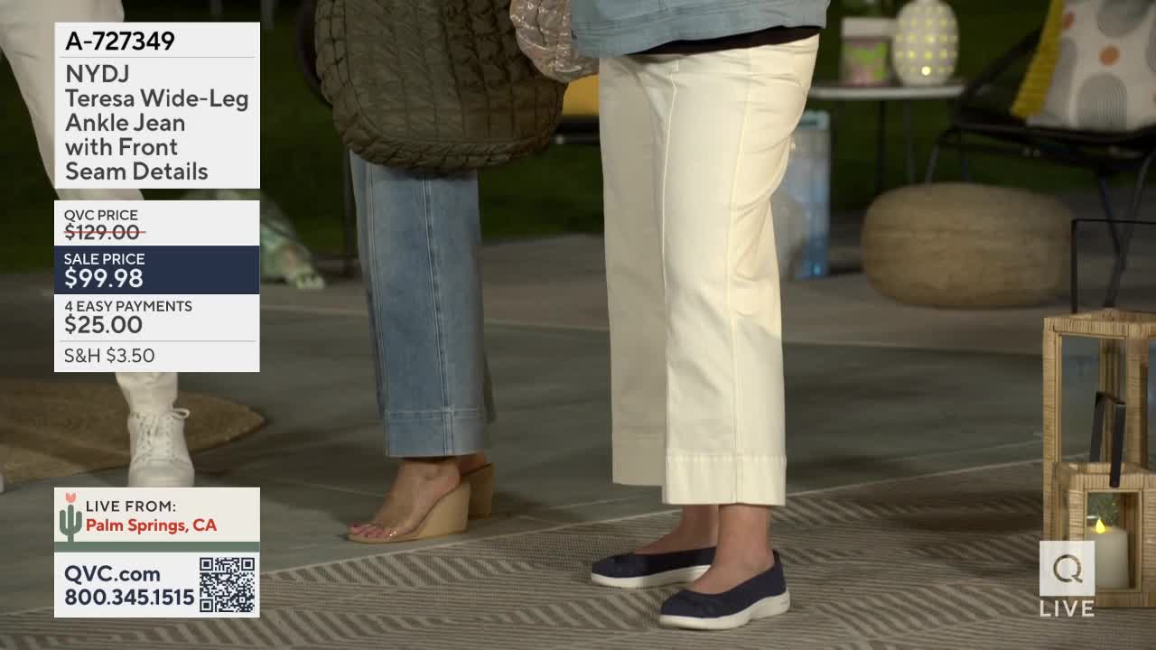 Two women stand on a patterned patio, one in light wash denim jeans and wedge sandals, the other in cream-colored pants and navy flats. A QVC graphic overlays the lower left corner, showing the item details and contact information.