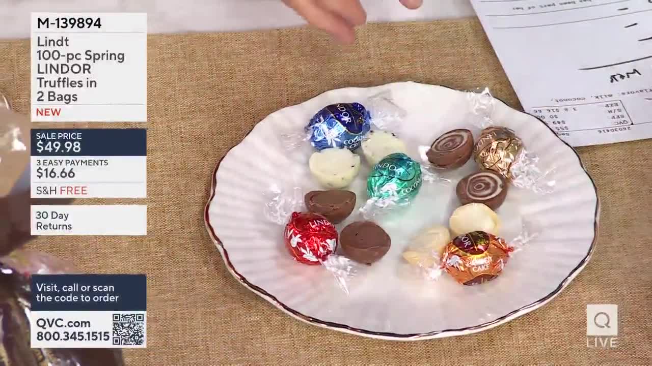 A hand reaches into the frame, hovering over a white, scalloped dish. The dish holds a collection of individually wrapped Lindt chocolates, their foil wrappers glinting under the lights.