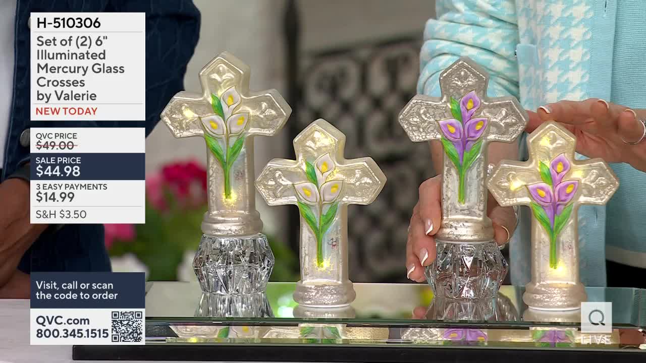 A hand reaches out to adjust a mercury glass cross, its delicate calla lilies illuminated from within. Another cross, identical but for its purple blooms, stands beside it.