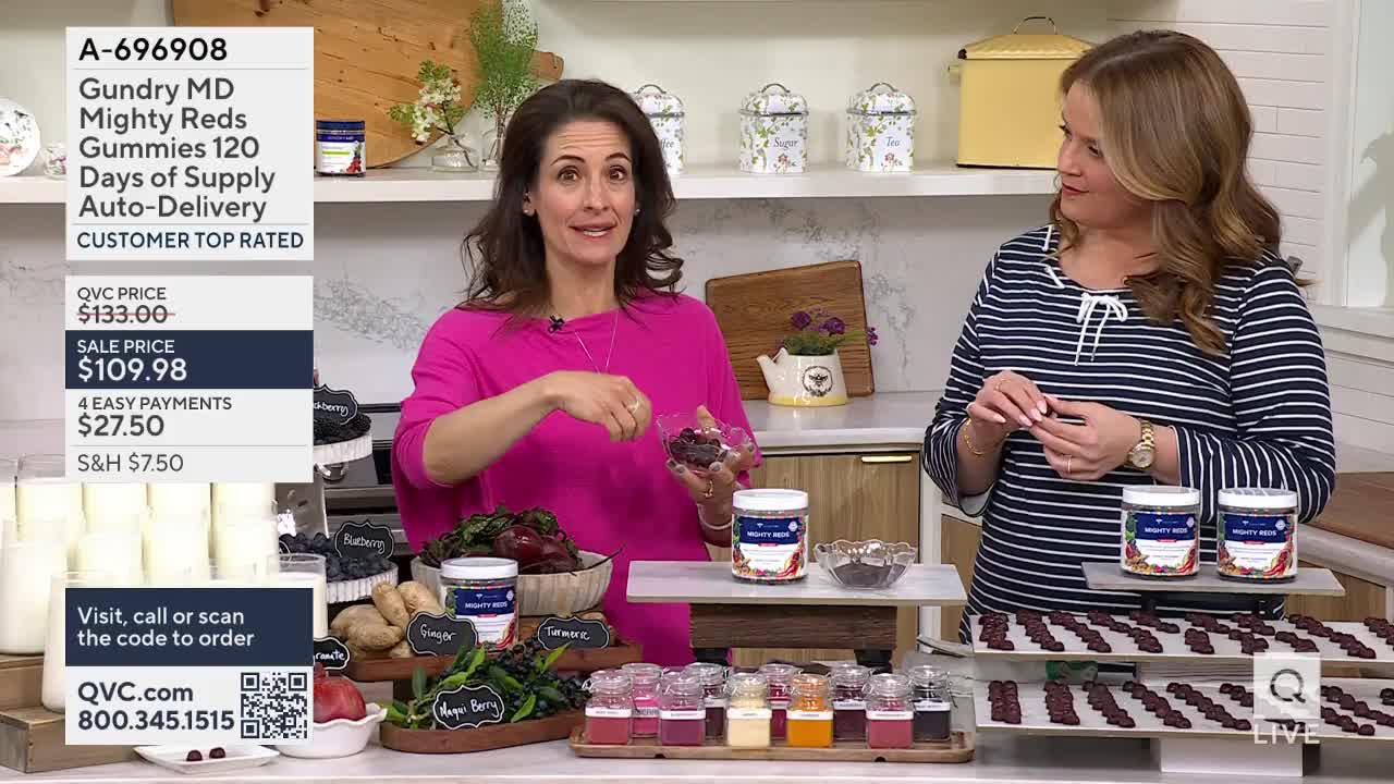 A woman in a bright pink top holds out a small dish of dark red gummies, her other hand gesturing towards a display of ingredients. Beside her, another woman in a striped shirt examines a gummy between her fingers.