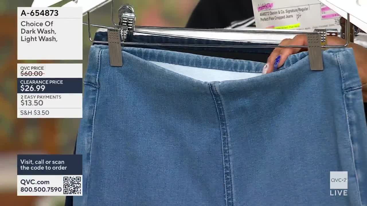 A hand adjusts a pair of light wash denim jeans hanging on a rack. The QVC2 logo is visible in the corner.
