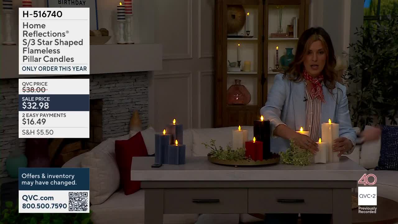 A woman on QVC2 arranges flameless pillar candles on a table. The candles glow with a warm, flickering light.