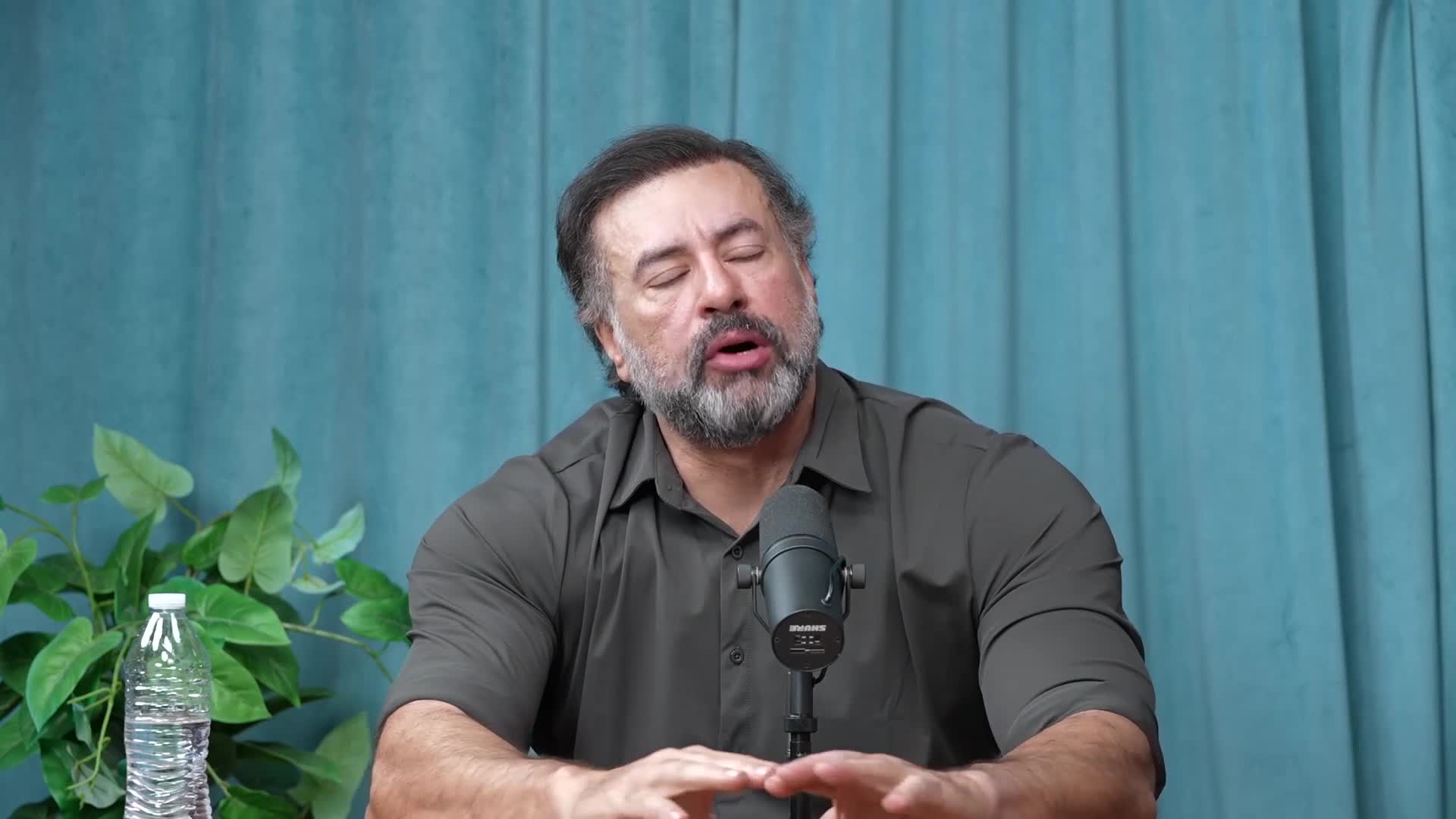 A man with a beard speaks into a microphone, his hands gesturing as he talks. A bottle of water and a leafy plant sit on a table beside him.