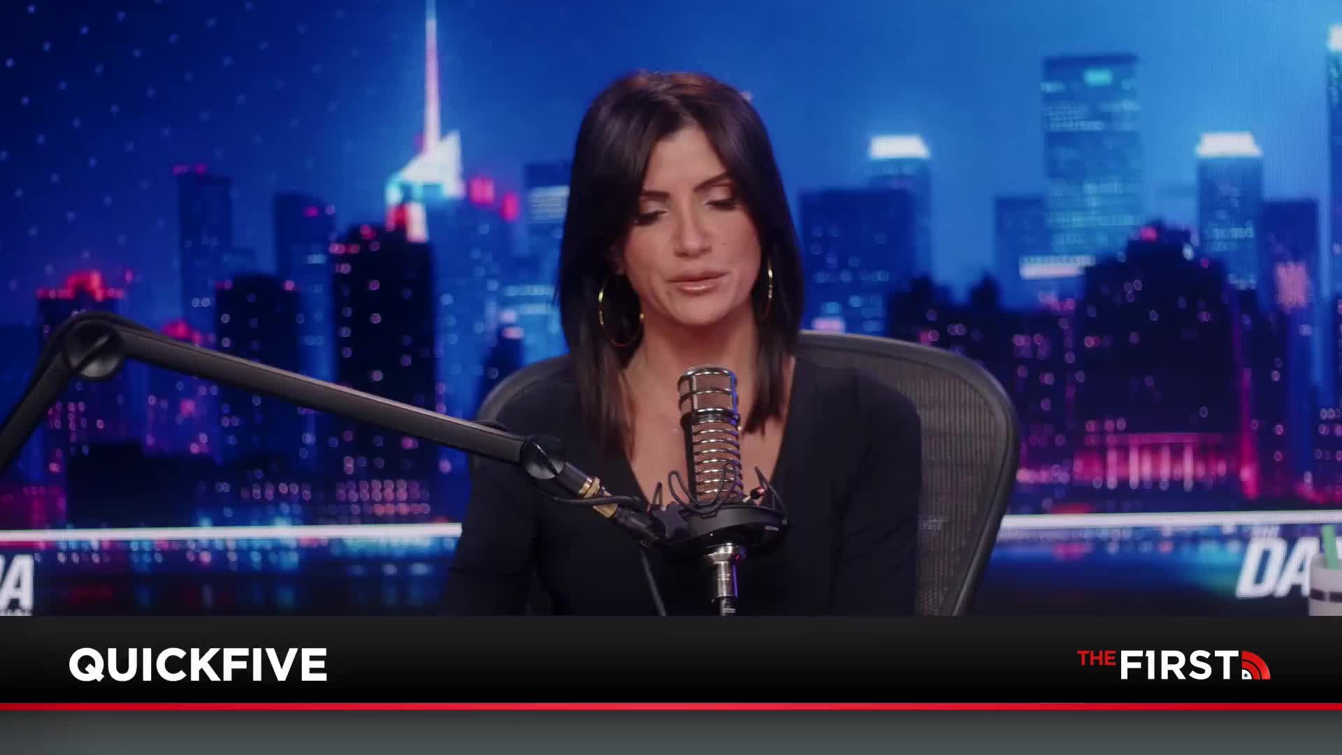 A woman with dark hair sits at a desk, speaking into a microphone. Behind her, a cityscape glows with neon lights.