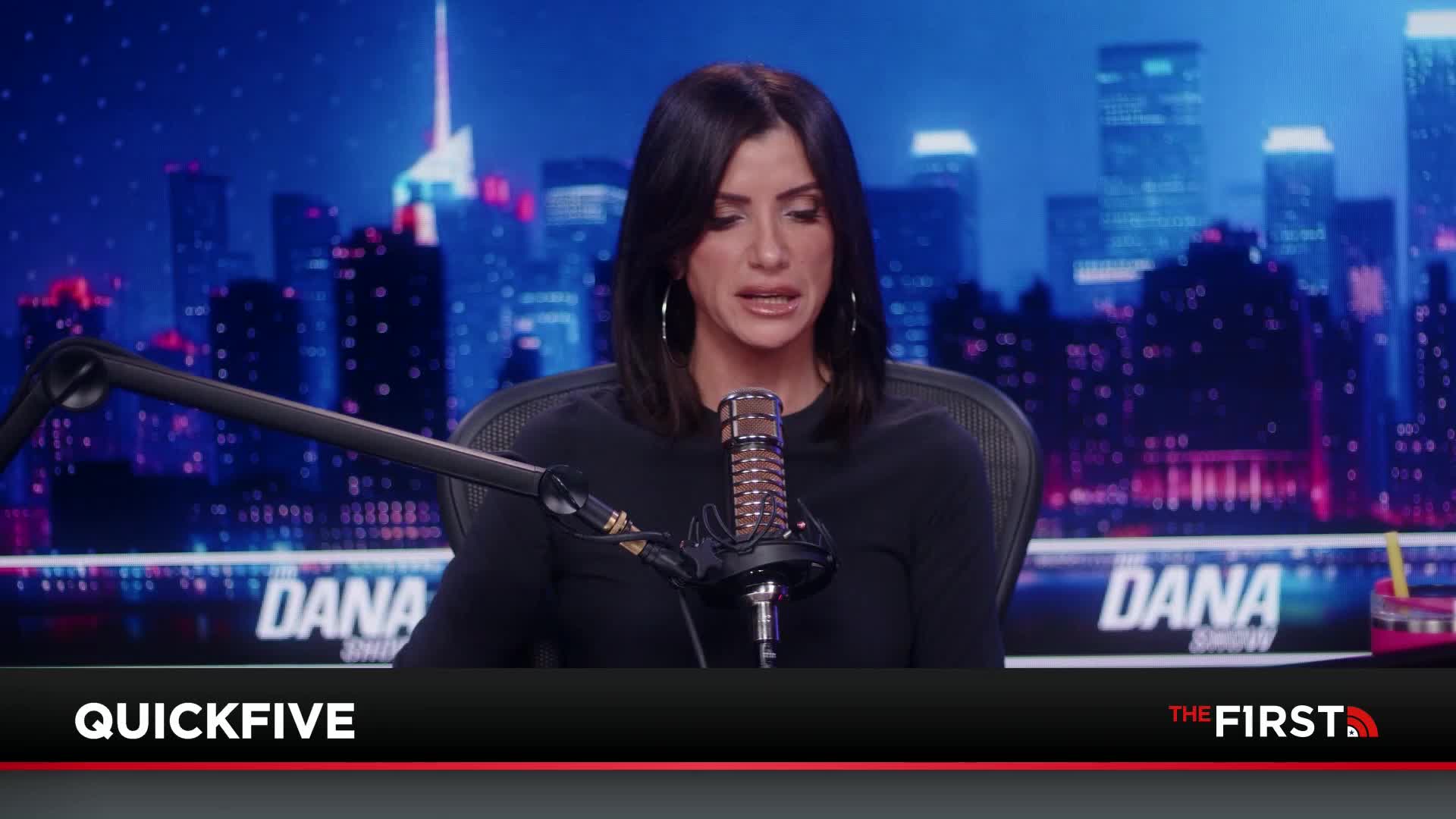 A woman with dark hair sits at a desk, speaking into a microphone. Behind her, a cityscape glows under a dark sky.