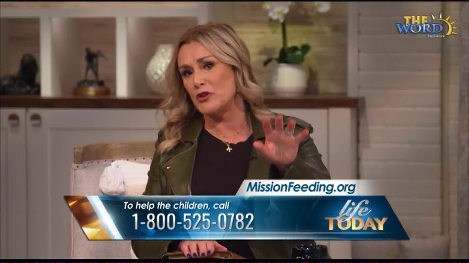 A woman with blonde hair, wearing a green jacket, gestures with her right hand as she speaks. A graphic overlays the bottom of the screen, displaying a phone number and website for MissionFeeding.org.