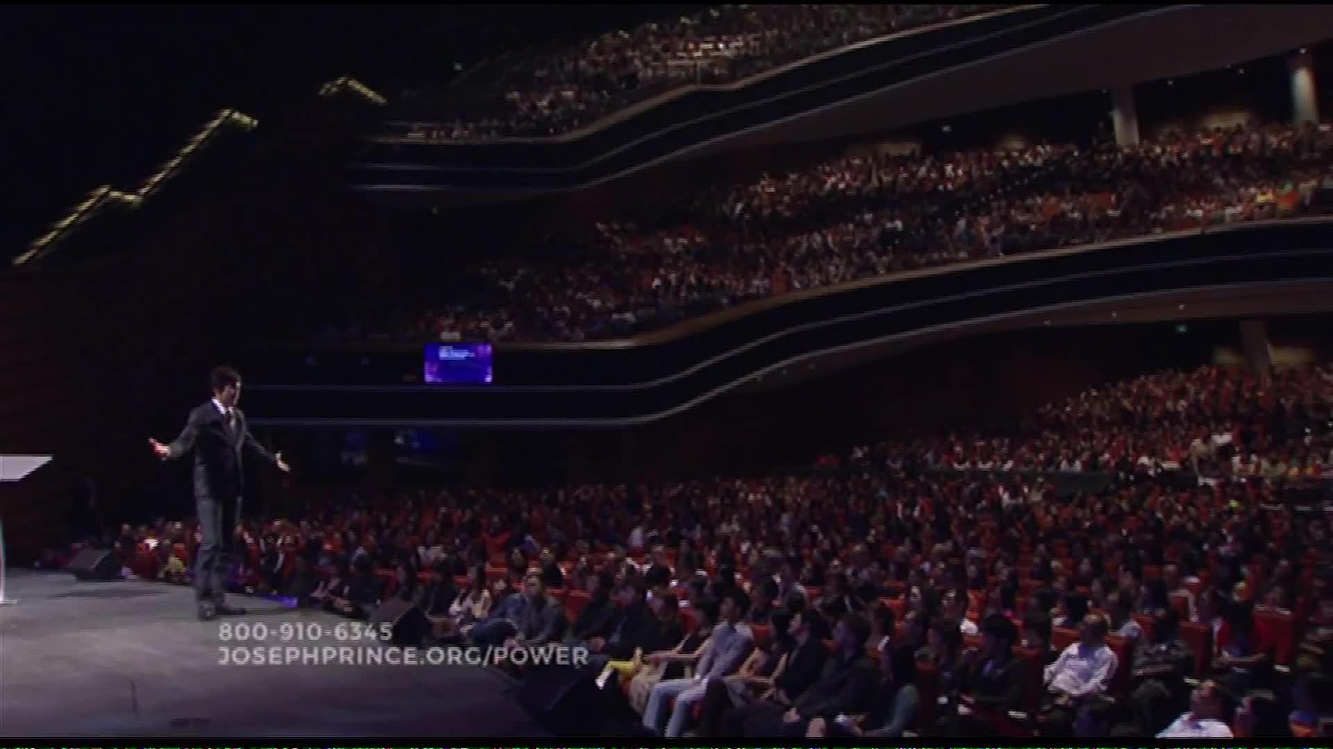 A man in a dark suit stands on a stage with his arms outstretched, addressing a massive crowd filling tiered seating. The Word Network broadcast shows the event from a wide angle, capturing the scale of the audience gathered in this United States venue.