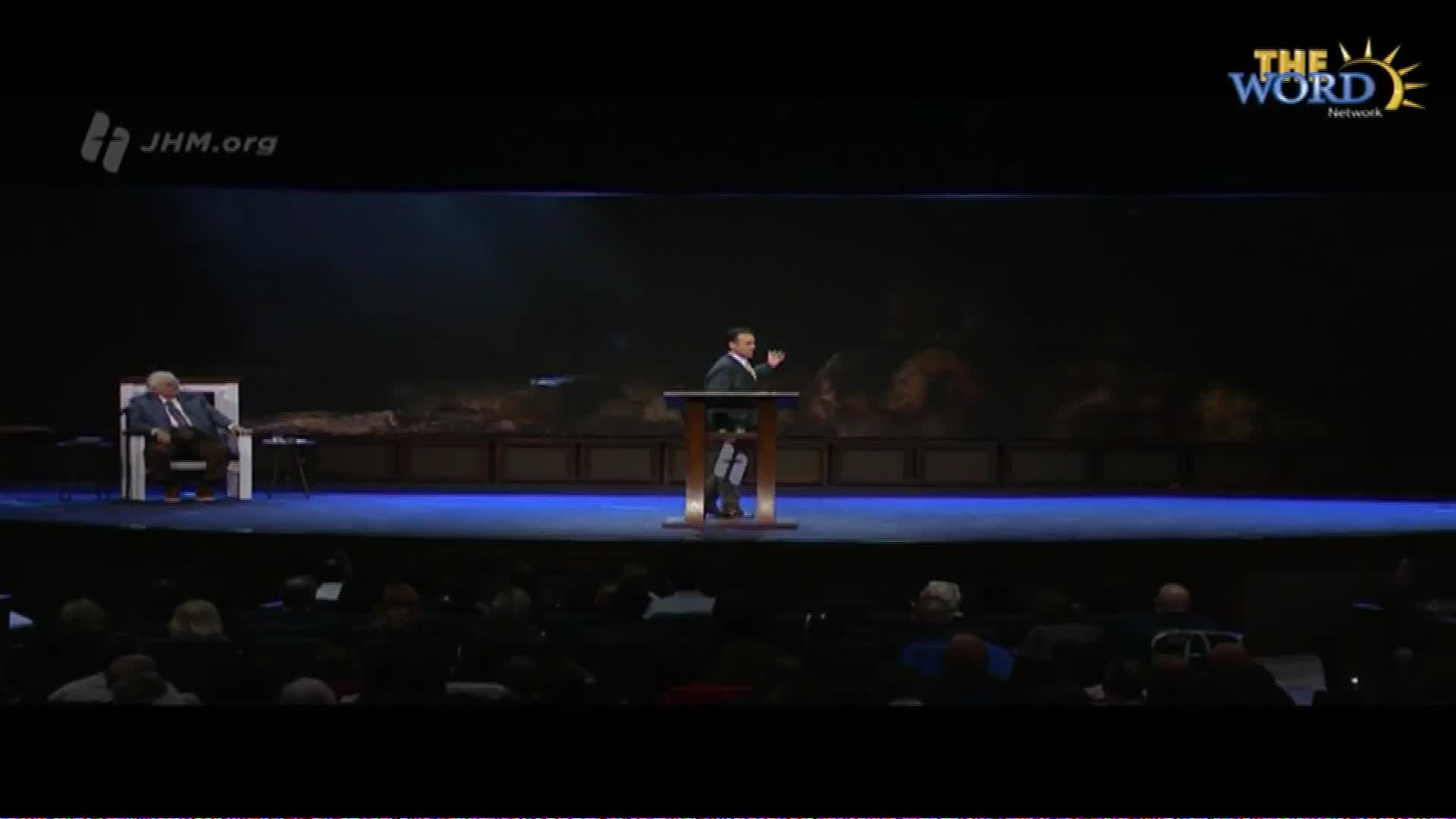 A man in a suit stands at a wooden podium, gesturing with his right hand. To his left, another man sits in a white chair. The Word Network logo appears in the upper right corner of the dark stage.