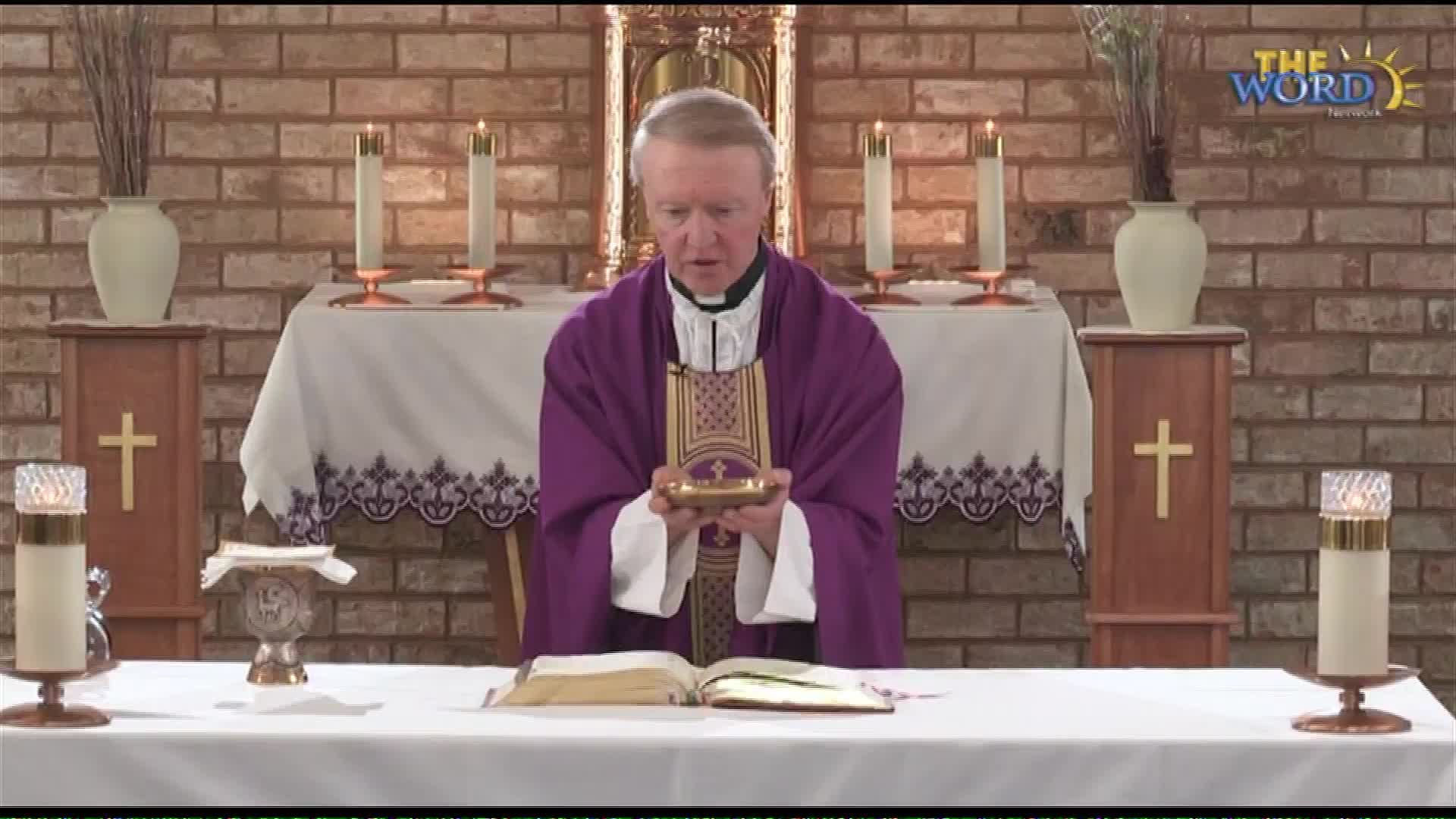 A priest in a purple vestment holds a golden chalice. He stands before an open book on a table, with lit candles flanking him.