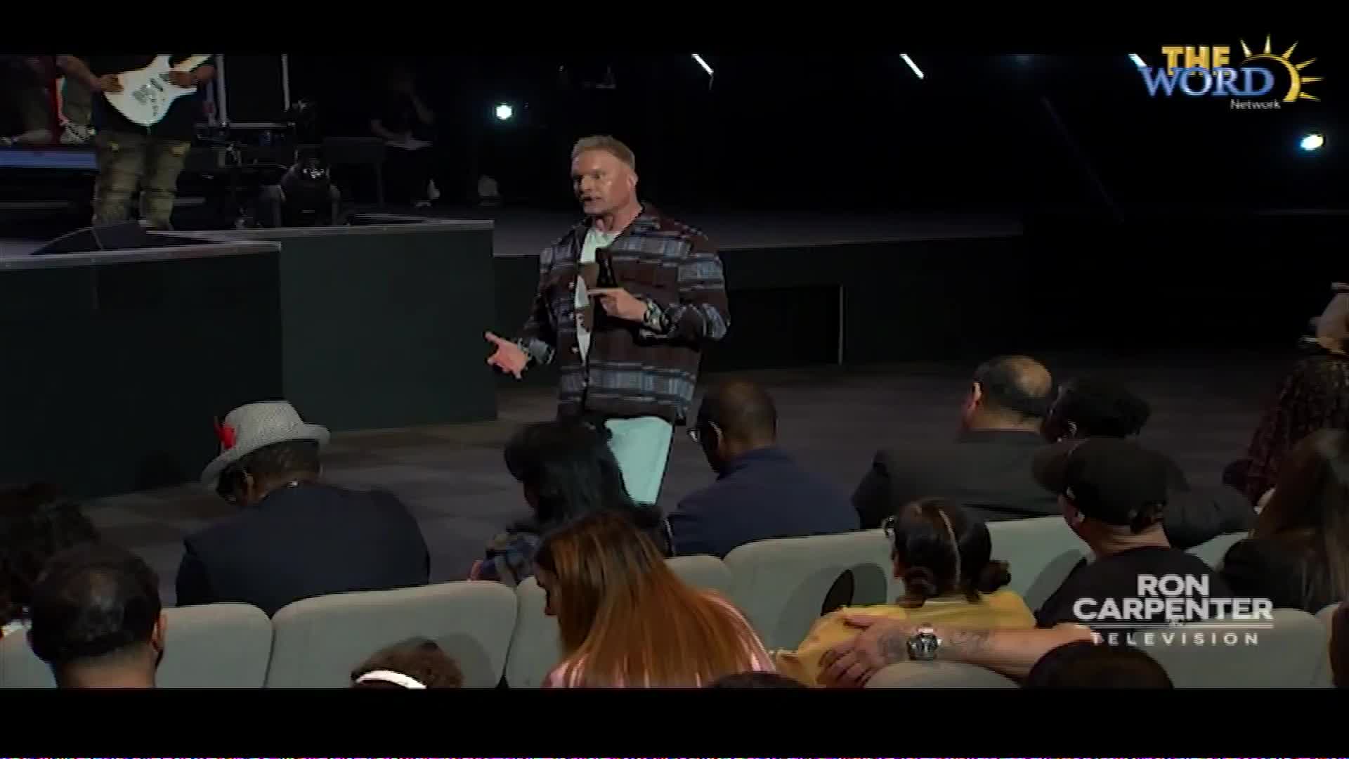 A man in a patterned shirt gestures with his hands as he speaks from a stage, while a guitarist stands behind him. The audience in front of the stage watches him intently.