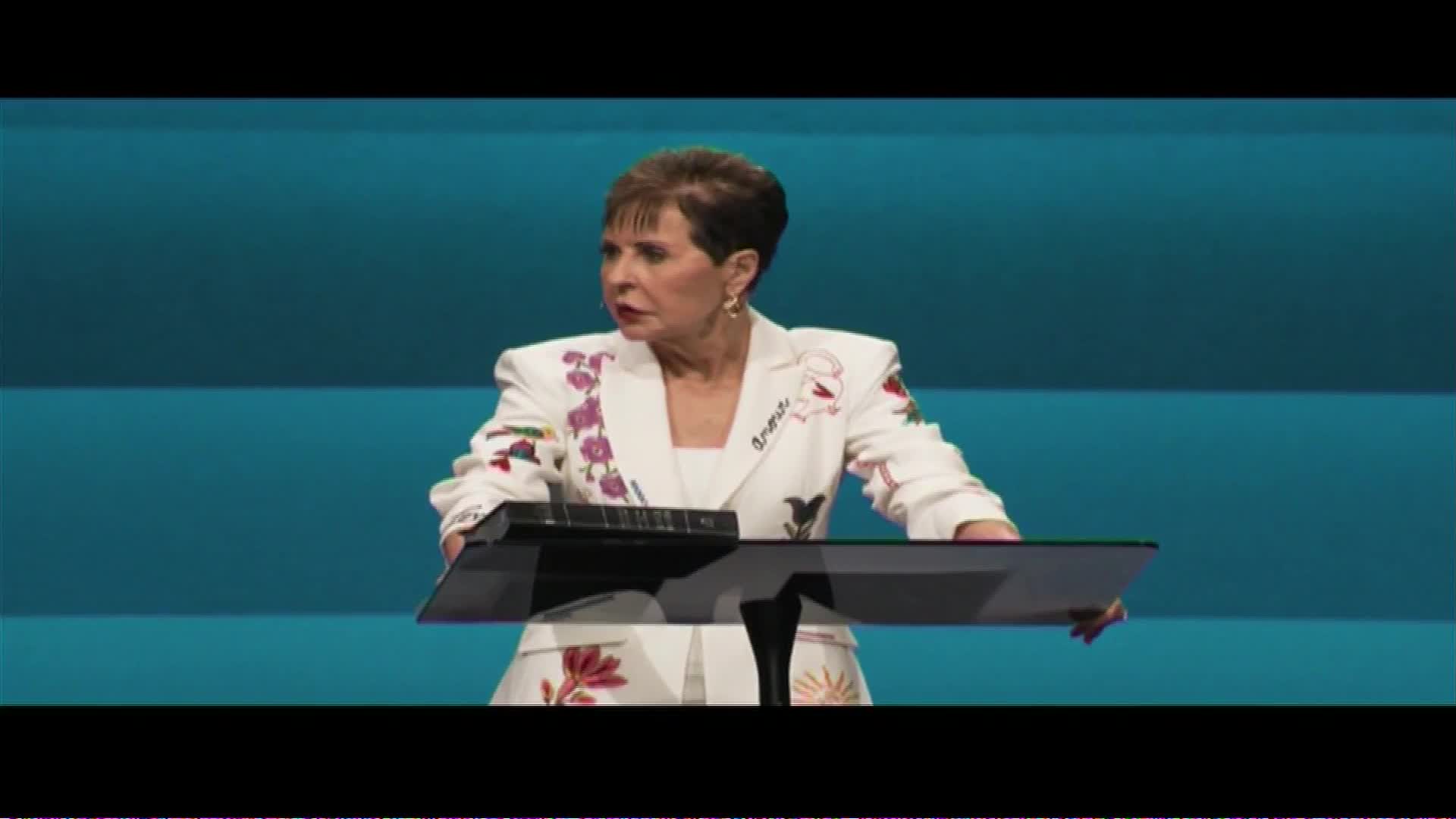 A woman in a white jacket with embroidered flowers stands behind a podium. She's speaking, her gaze directed off to the side. This is likely a broadcast from The Word Network.