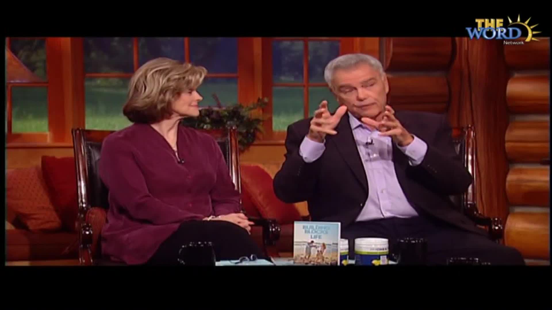 A man gestures with his hands, explaining something on The Word Network. A woman sits beside him, listening intently.