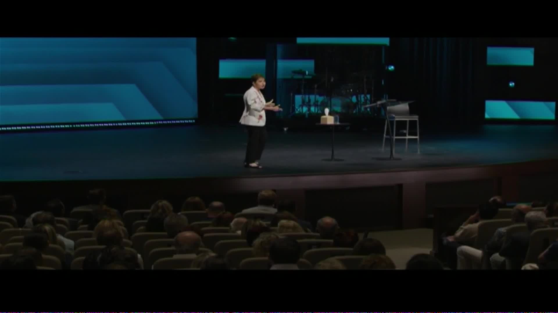 A woman stands on a stage, speaking to an audience. The stage is lit, with a large screen behind her displaying abstract blue shapes.