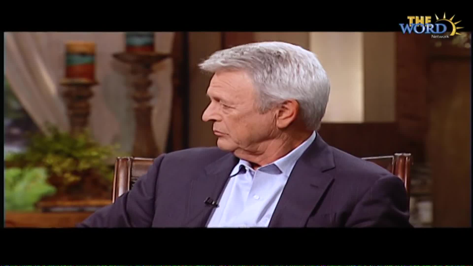 A man with silver hair, wearing a suit jacket over a light blue shirt, is seated and looking off to the side. The logo for The Word Network appears in the upper right corner of the screen.