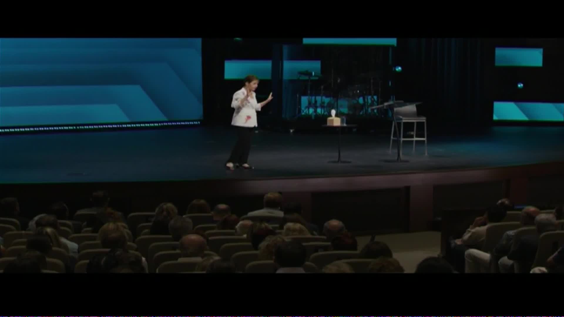 A woman stands on a stage, gesturing with her hands. An audience sits in rows, facing the stage.