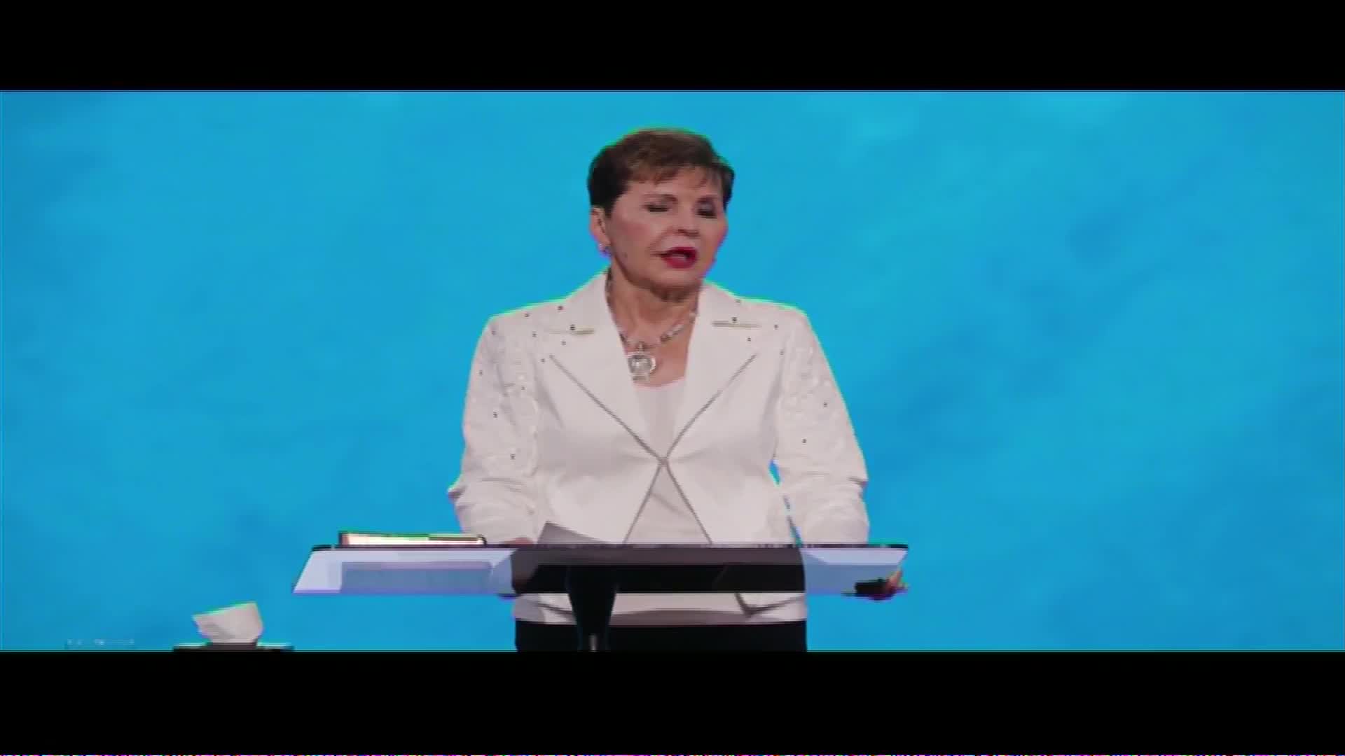 A woman stands behind a lectern, speaking into a microphone. She wears a white jacket with decorative studs, and a bright blue backdrop fills the space behind her.