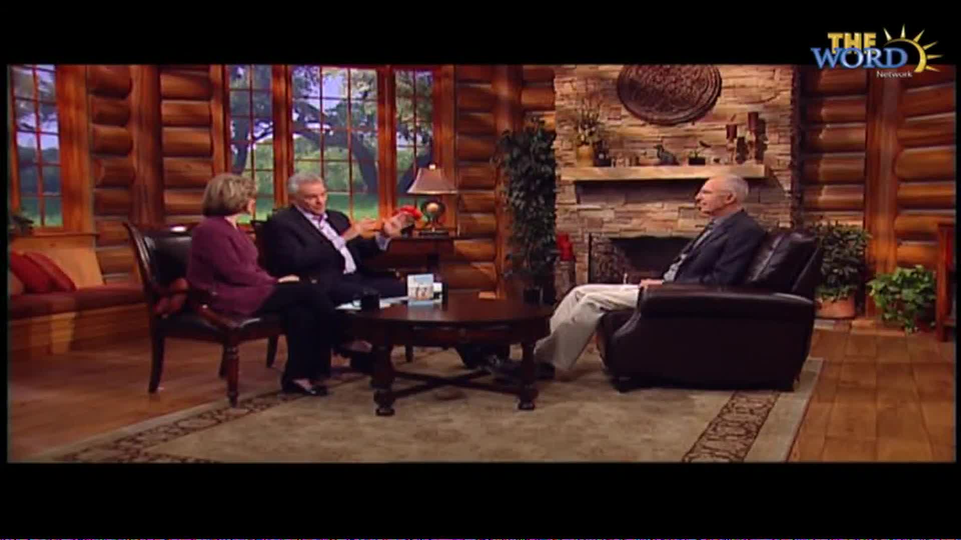 A man in a suit gestures with his hands while speaking to a woman seated across from him. Another man sits in a recliner to the right, listening intently. The scene unfolds in a cozy, log-cabin-style studio, likely for a program on The Word Network.