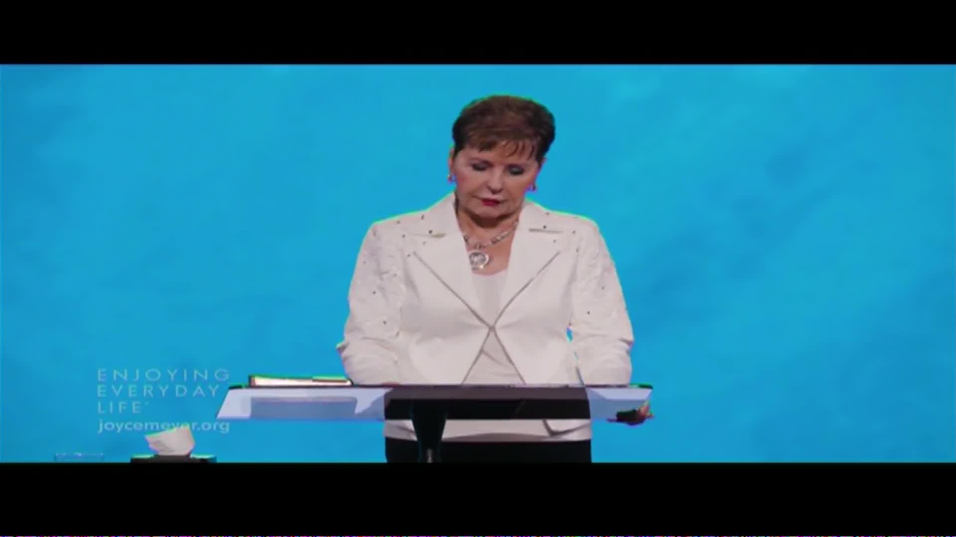 Joyce Meyer stands behind a podium, her gaze lowered as if in thought. The backdrop is a solid, bright blue, with "ENJOYING EVERYDAY LIFE" and "joycemeyer.org" subtly displayed to the left.