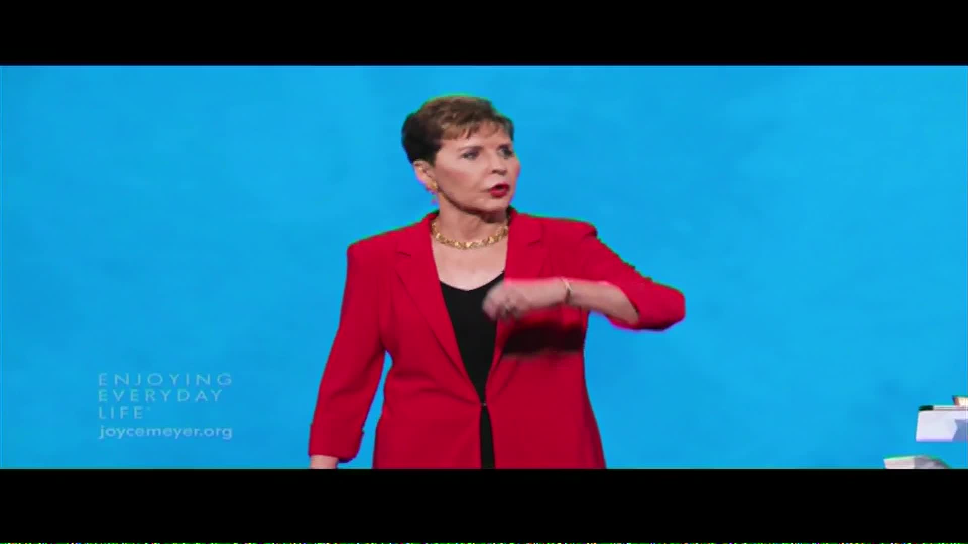 A woman in a bright red blazer gestures with her right hand. She speaks from a stage with a vibrant blue backdrop, likely for a broadcast on The Word Network.