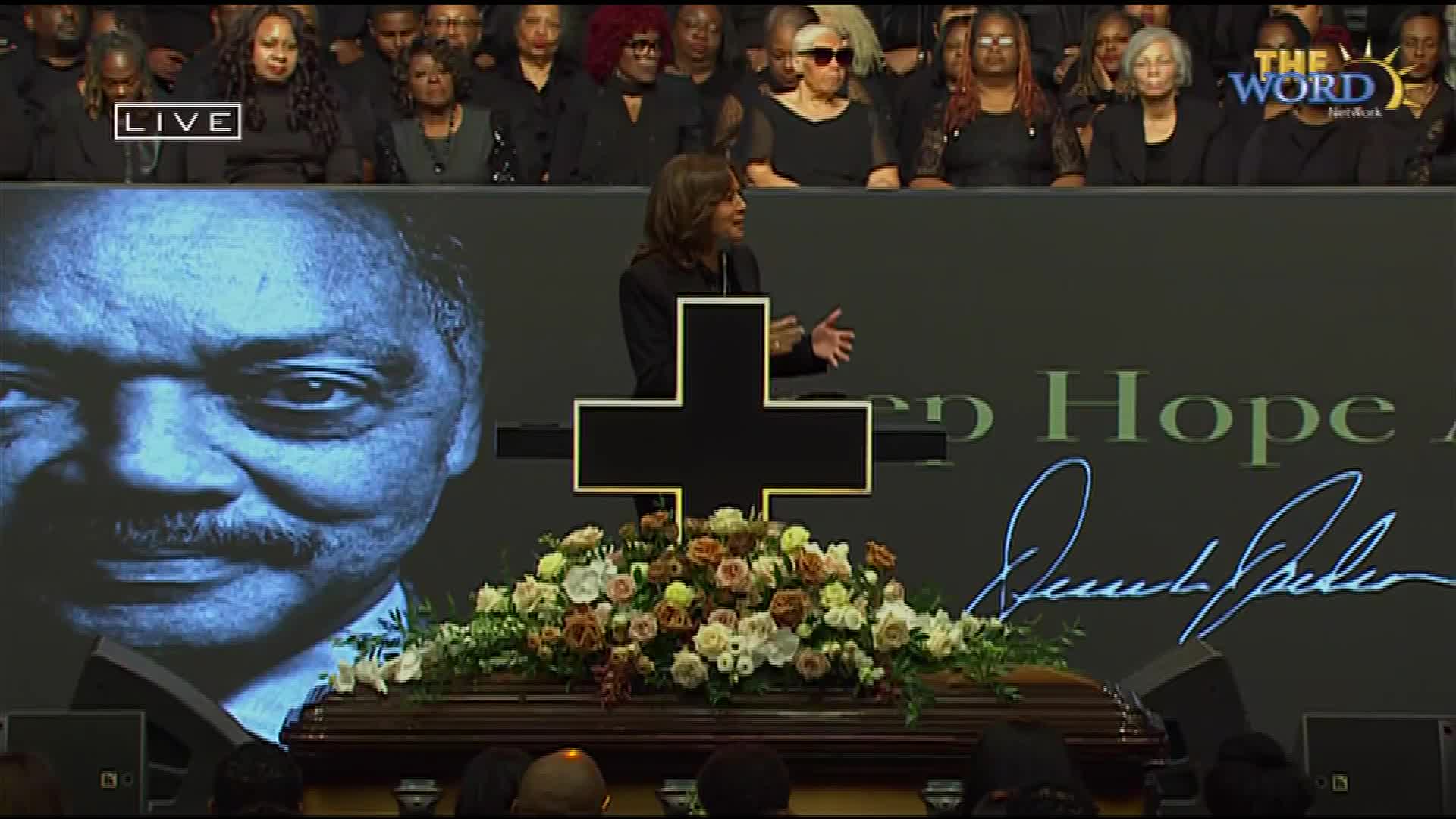 A woman speaks from behind a podium shaped like a cross, her hands gesturing. Before her, a casket rests, adorned with a large floral arrangement.