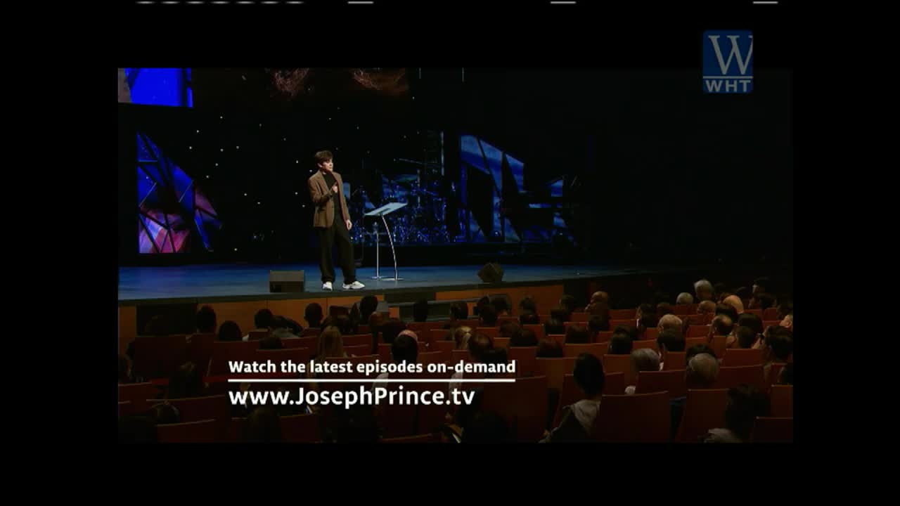 A man in a brown jacket stands on a stage, speaking into a microphone. The audience is seated, watching the broadcast from World Harvest TV.
