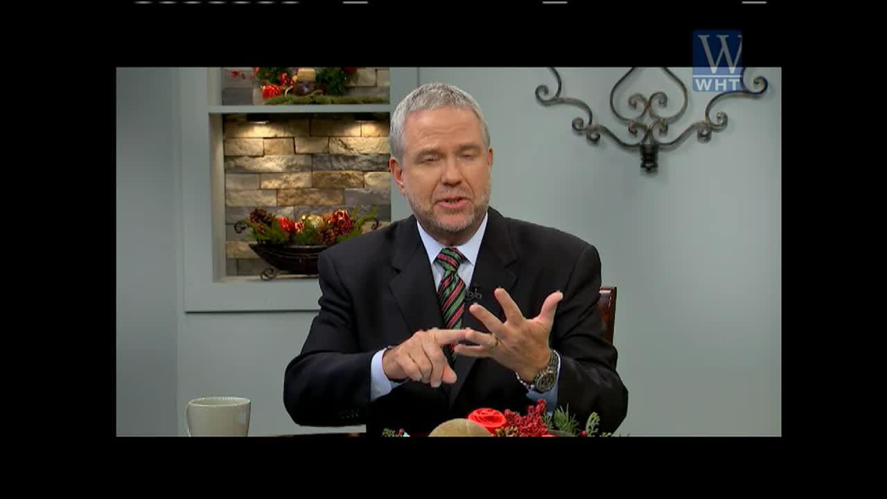He's gesturing with his right hand, counting off points as he speaks, while his left hand rests on the table. The man in the suit is on World Harvest TV, the WHT logo is in the upper right.
