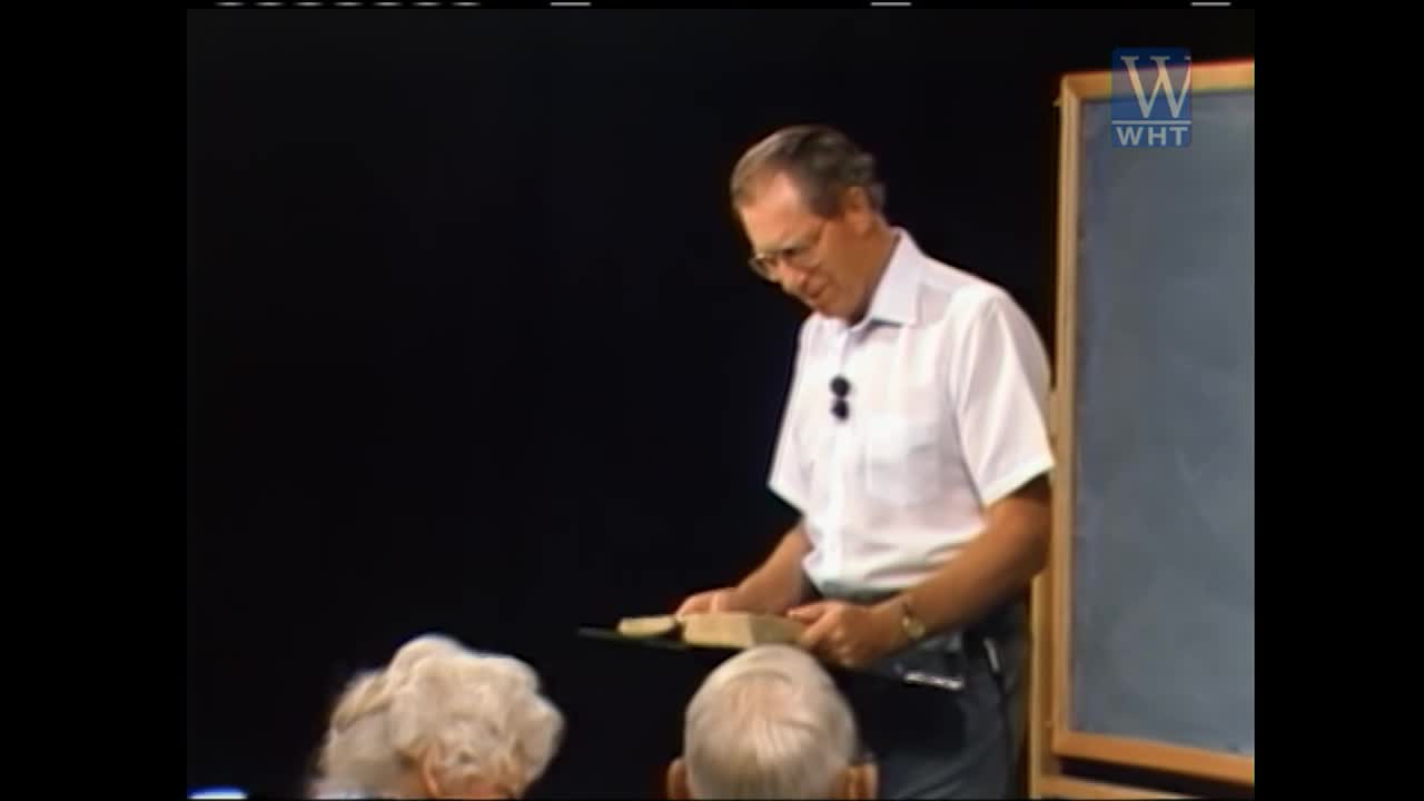 A man in a white shirt stands at a lectern, holding an open book. He looks down at the pages, his glasses reflecting the light. Behind him, a chalkboard with the World Harvest TV logo is visible.
