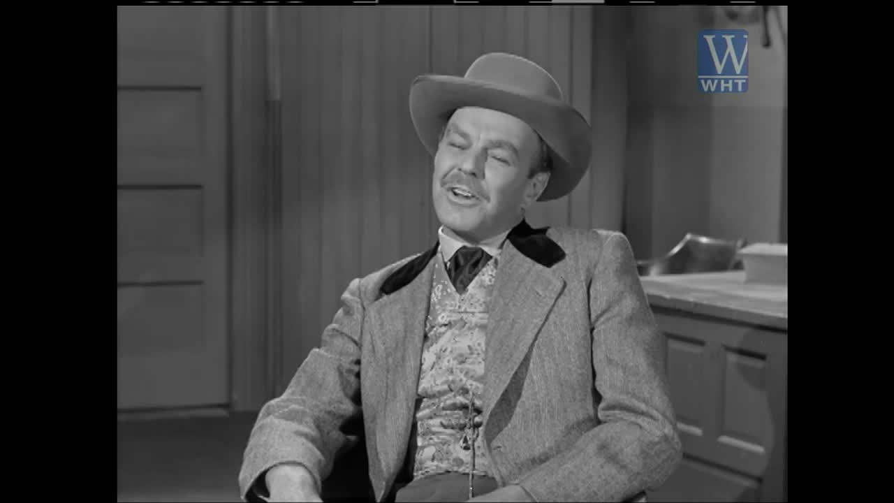 A man in a cowboy hat and vest leans back in his chair, eyes closed as he speaks. He's wearing a light-colored jacket over a patterned vest, and a dark tie.