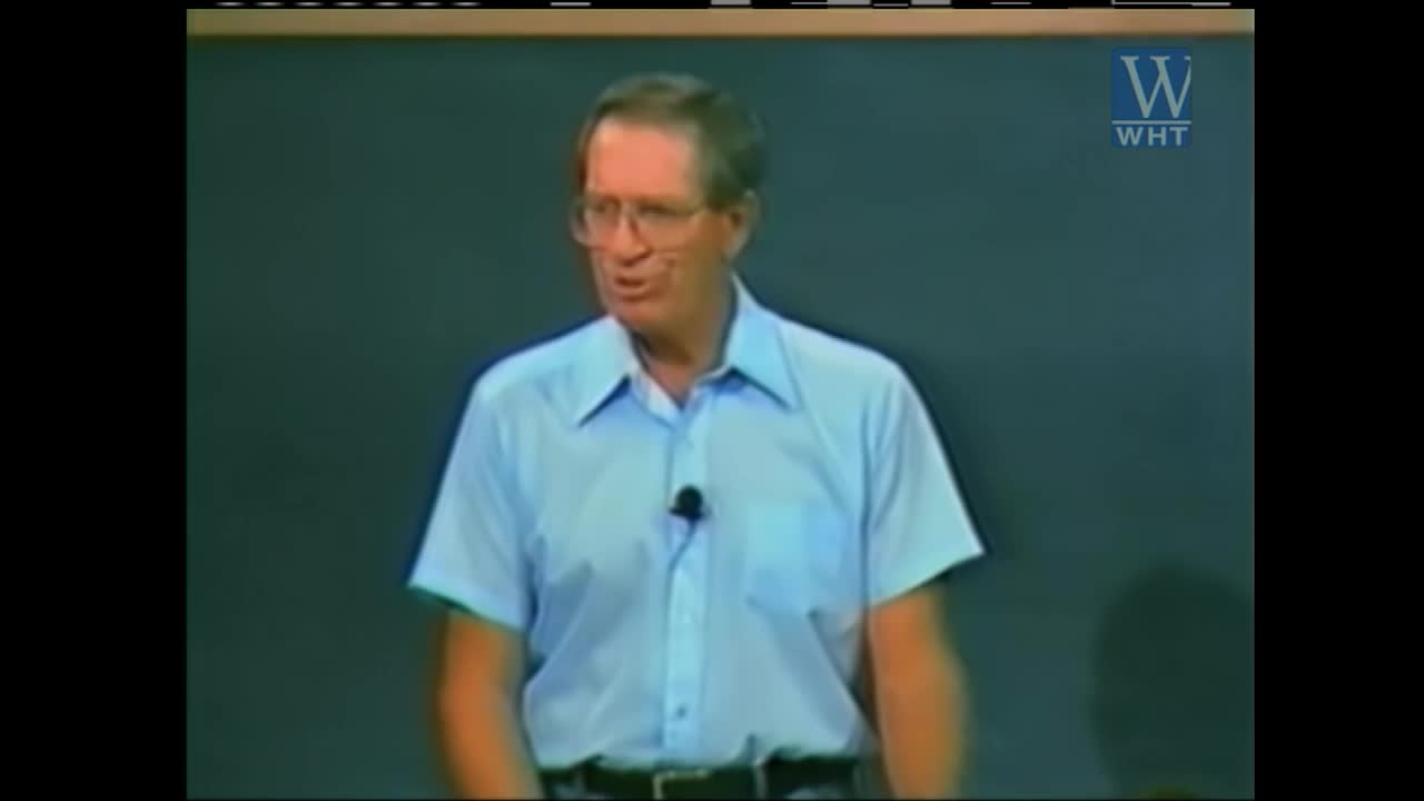 A man in a light blue collared shirt stands before a dark green backdrop, speaking into a microphone clipped to his collar. A "WHT" logo appears in the upper right corner.