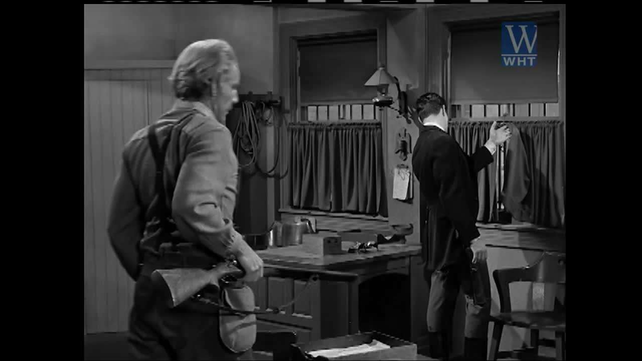 A man in overalls, a rifle slung low, turns from a workbench. Another man in a suit hangs a jacket behind a curtain.