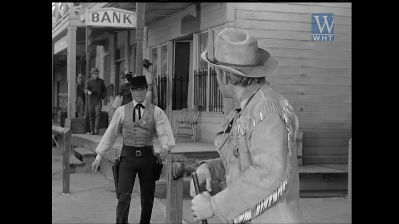 A man in a cowboy hat and fringed jacket stands facing a man in a vest and hat, both holding canes. The bank looms behind them, a sign of the United States' growing settlements.