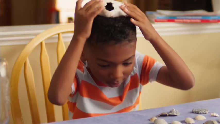 A young boy, wearing a striped shirt, is holding a deflated soccer ball above his head. He's looking down at a collection of seashells on the table in front of him.
