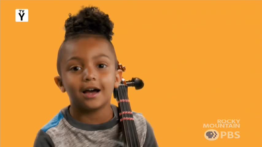 A young boy with a high ponytail stands holding a cello. He looks directly at me, his mouth open as if speaking.