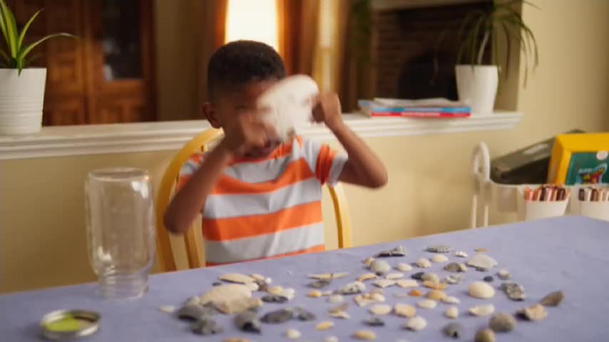 The boy is holding a large shell, shaking it as if to get something out. He's sitting at a table covered in smaller shells, with a glass jar and a small tin nearby.