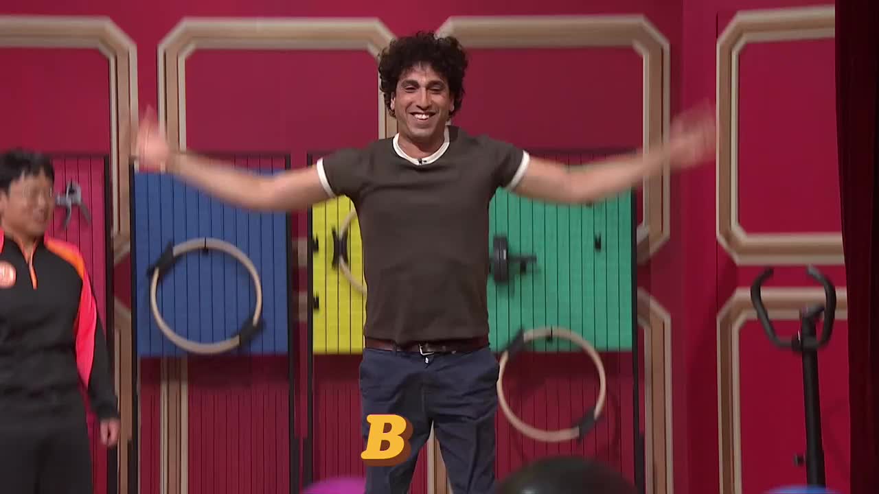 A man with curly hair is smiling, arms outstretched, in front of a colorful backdrop. Someone in a track suit stands to his left, watching.
