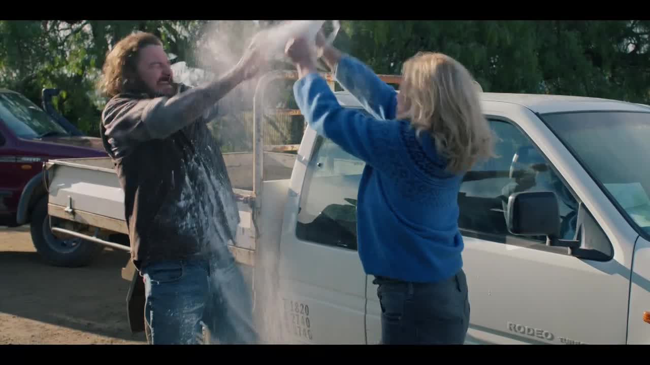 A woman in a blue sweater is pouring something white over a man standing next to a white ute. The man is getting drenched, and the white substance is clouding the air between them.
