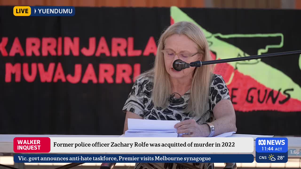 A woman with blonde hair and glasses reads from a stack of papers at a podium. Behind her, a banner with bold red text and a green gun graphic is visible.
