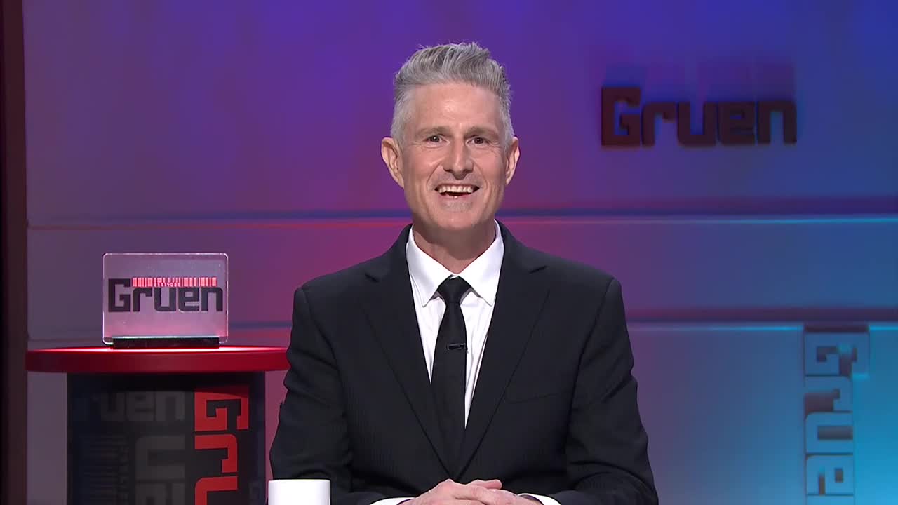 A man in a black suit and tie smiles at the camera, standing in front of a "Gruen" sign. The set on ABC Australia is lit with purple and blue hues.
