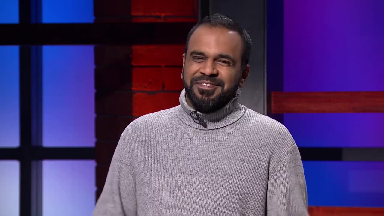 A man with a beard and a grey turtleneck smiles, looking directly at the camera. Behind him, the set has a brick wall and a purple backdrop.
